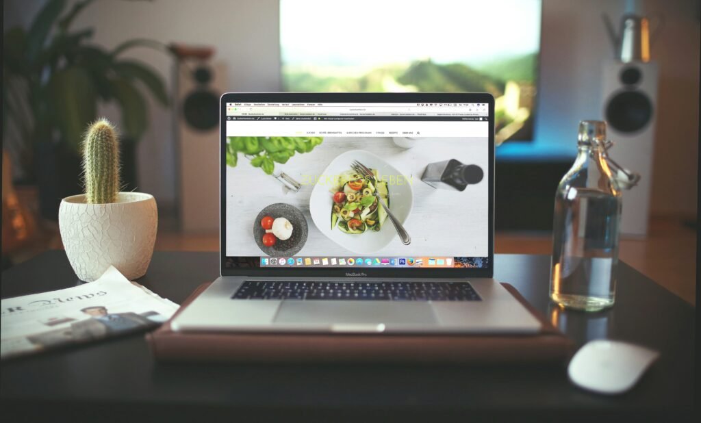 Laptop open on a desk showing a food recipe website, next to a cactus plant and a glass water bottle